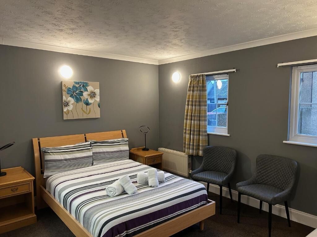 Una camera da letto con un letto, due sedie, un comodino, un termosifone, una finestra con tende e un quadro appeso al muro.