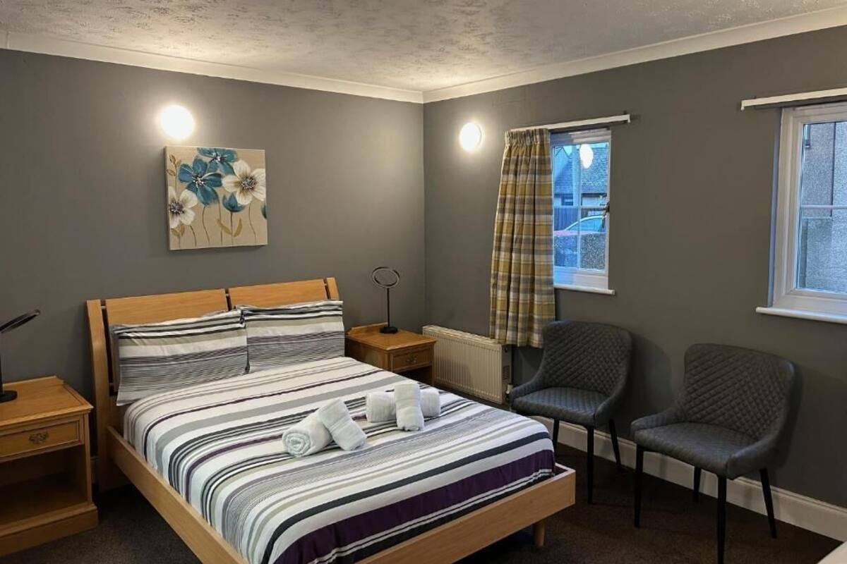 A bedroom with a bed, two chairs, a nightstand, a radiator, a window with curtains, and a painting on the wall.