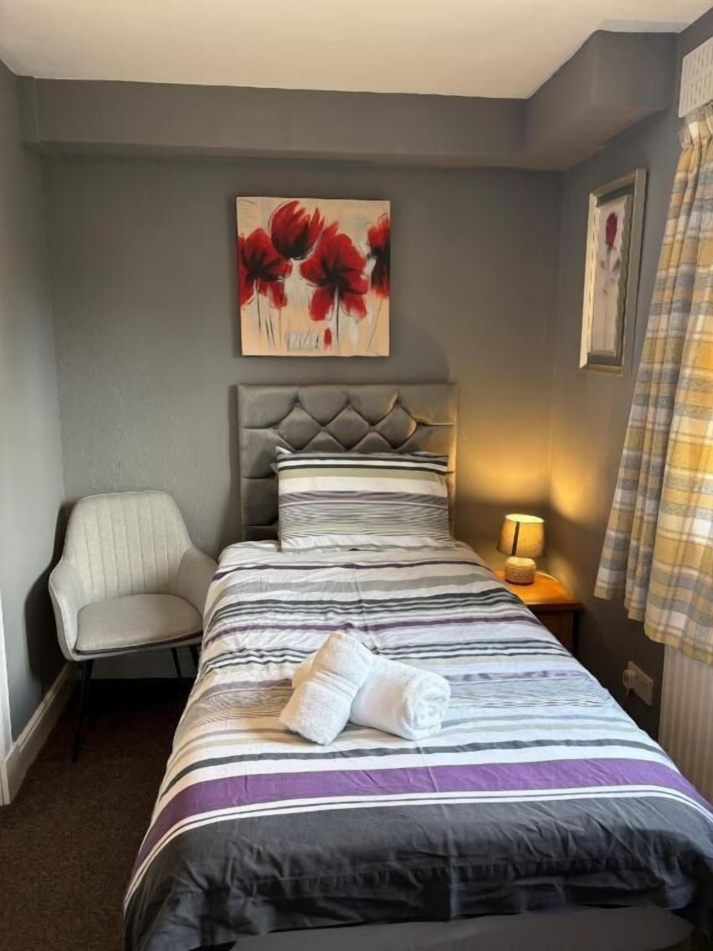 Una camera d'albergo con un letto, una sedia, un quadro, una lampada e una finestra con tende.