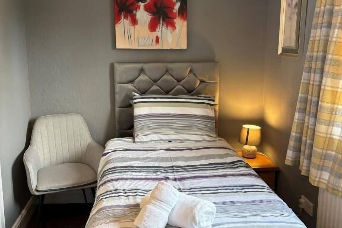 A hotel room with a bed, a chair, a painting, a lamp, and a window with curtains.