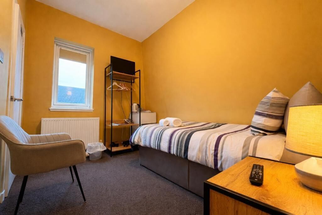 Camera d'albergo compatta con un letto singolo, una sedia, un comodino con telecomando e una finestra con persiane.