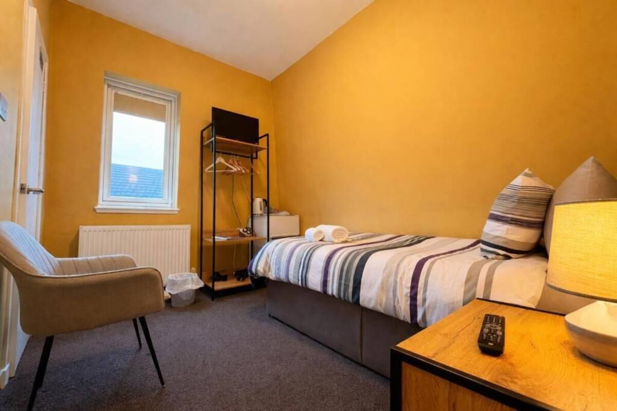 A compact hotel room with a single bed, a chair, a bedside table with a remote, and a window with blinds.