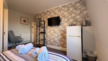 A hotel room with a bed, a chair, a TV, a refrigerator, and a trash bin.