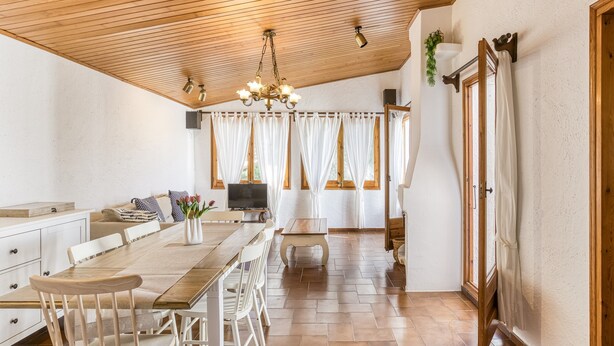 A spacious kitchen with wooden flooring, a long dining table, white chairs, a chandelier, and large windows with curtains.