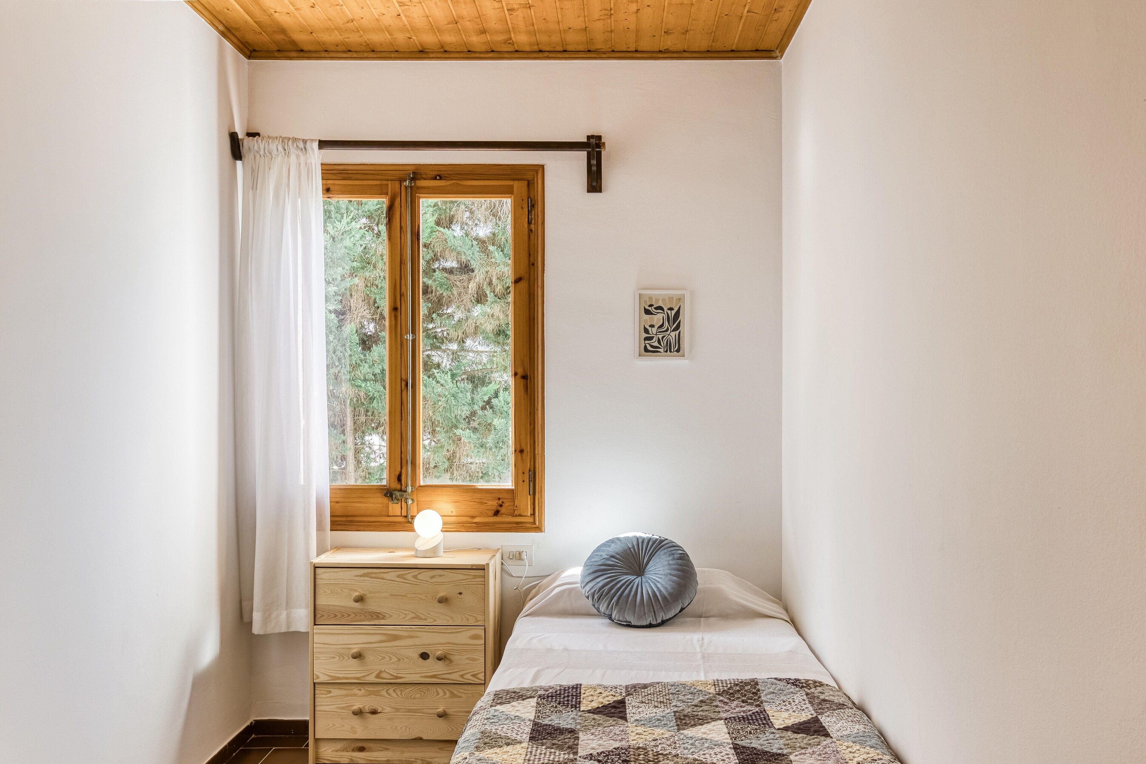 Een slaapkamer met een houten plafond, een raam met gordijn, een nachtkastje met lamp, een bed met kussen, en een aan de muur hangend schilderij.