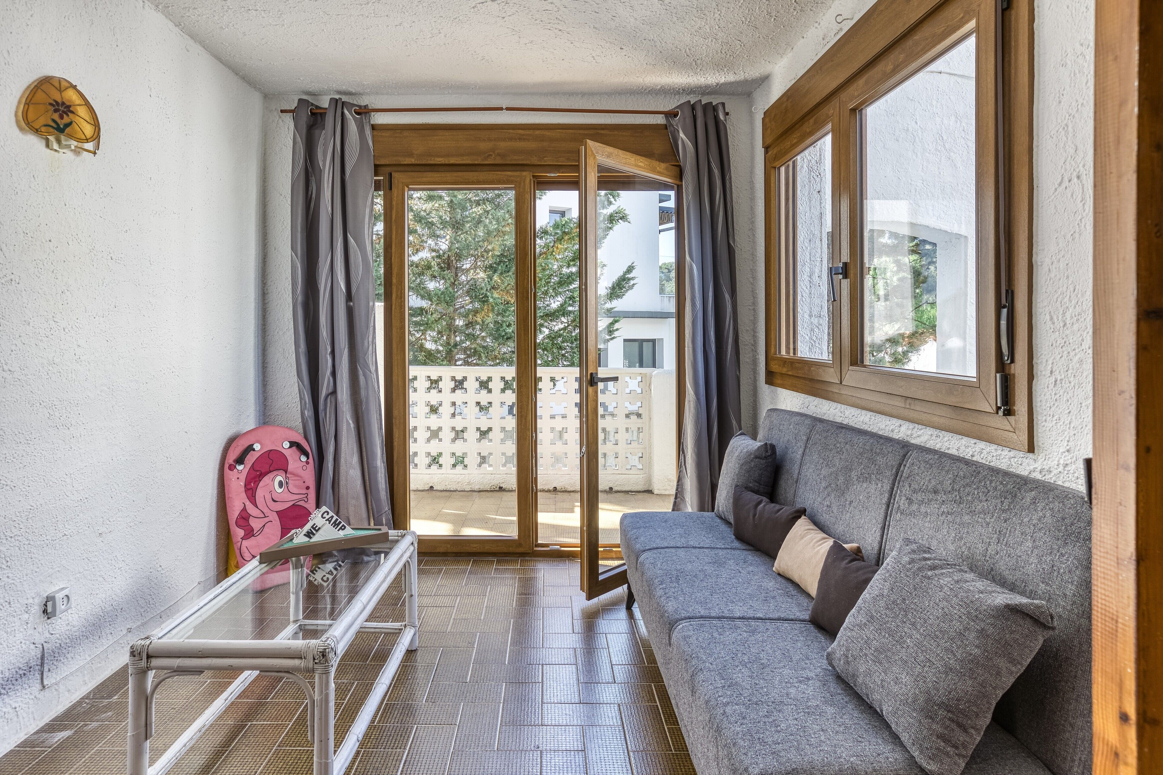 Een kamer met een grijze bank, een glazen tafel en een schuifpui naar een buitenruimte.