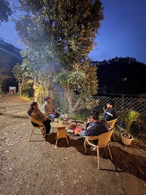 Een groep mensen zit rond een vuurkuil, omringd door rieten stoelen en potplanten, in een buitengebied met bomen en een hek.