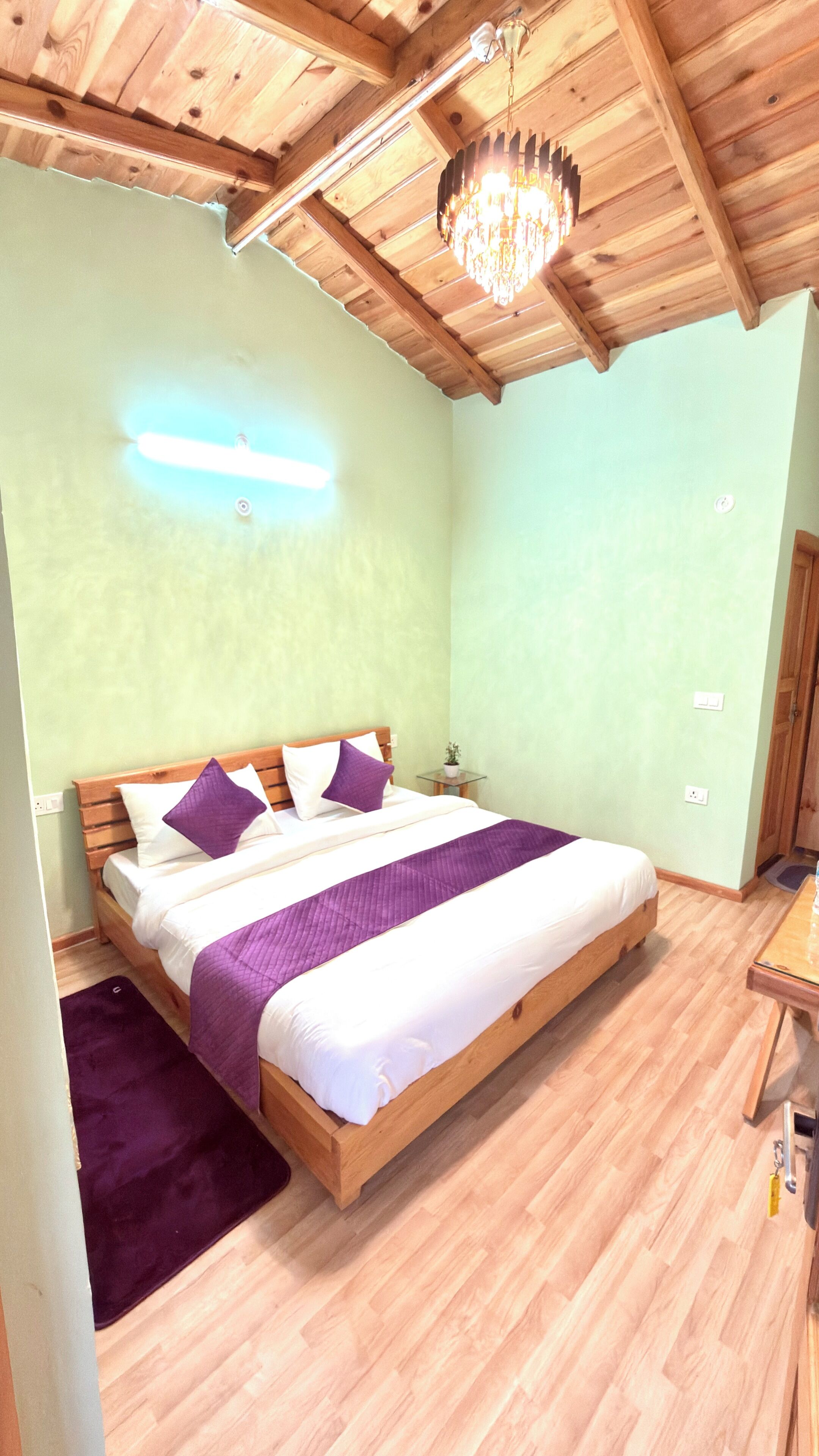 A bedroom with a wooden bed, a nightstand, a small table, a chandelier, and a wooden ceiling.