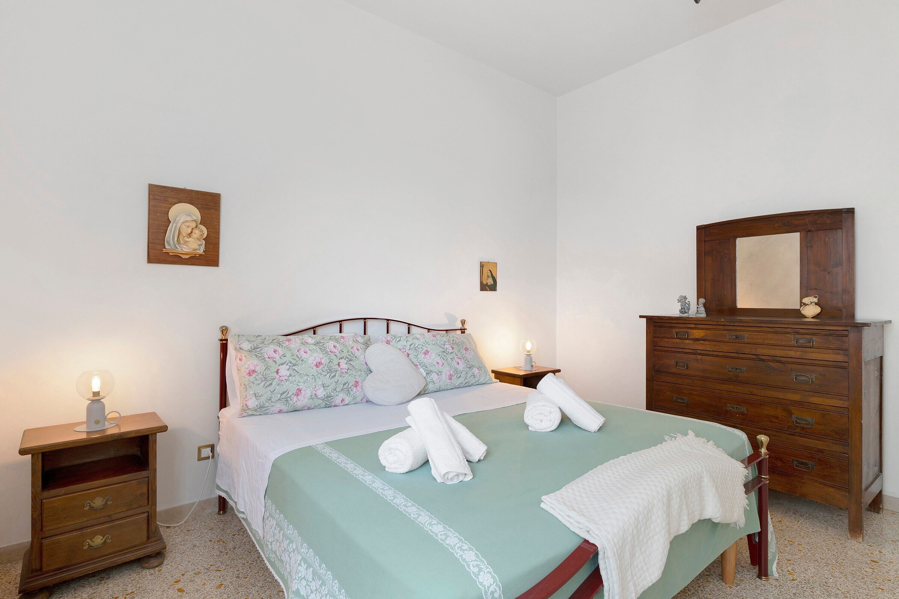 Une chambre à coucher comprenant un lit, une commode en bois, une table de chevet et une lampe fixée au mur.