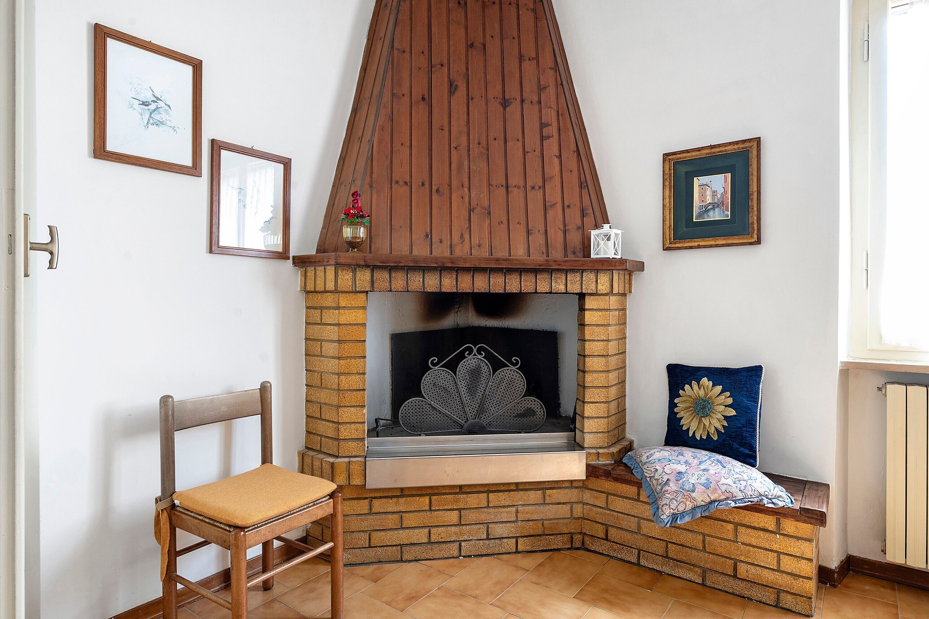 A corner fireplace with a brick surround, a wooden chair, and two framed pictures on the wall.