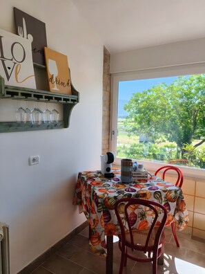 Een eethoek met een tafel waarop een tafelkleed met pompoenmotieven ligt, rode stoelen, een koffiezetapparaat en uitzicht op groen buiten.