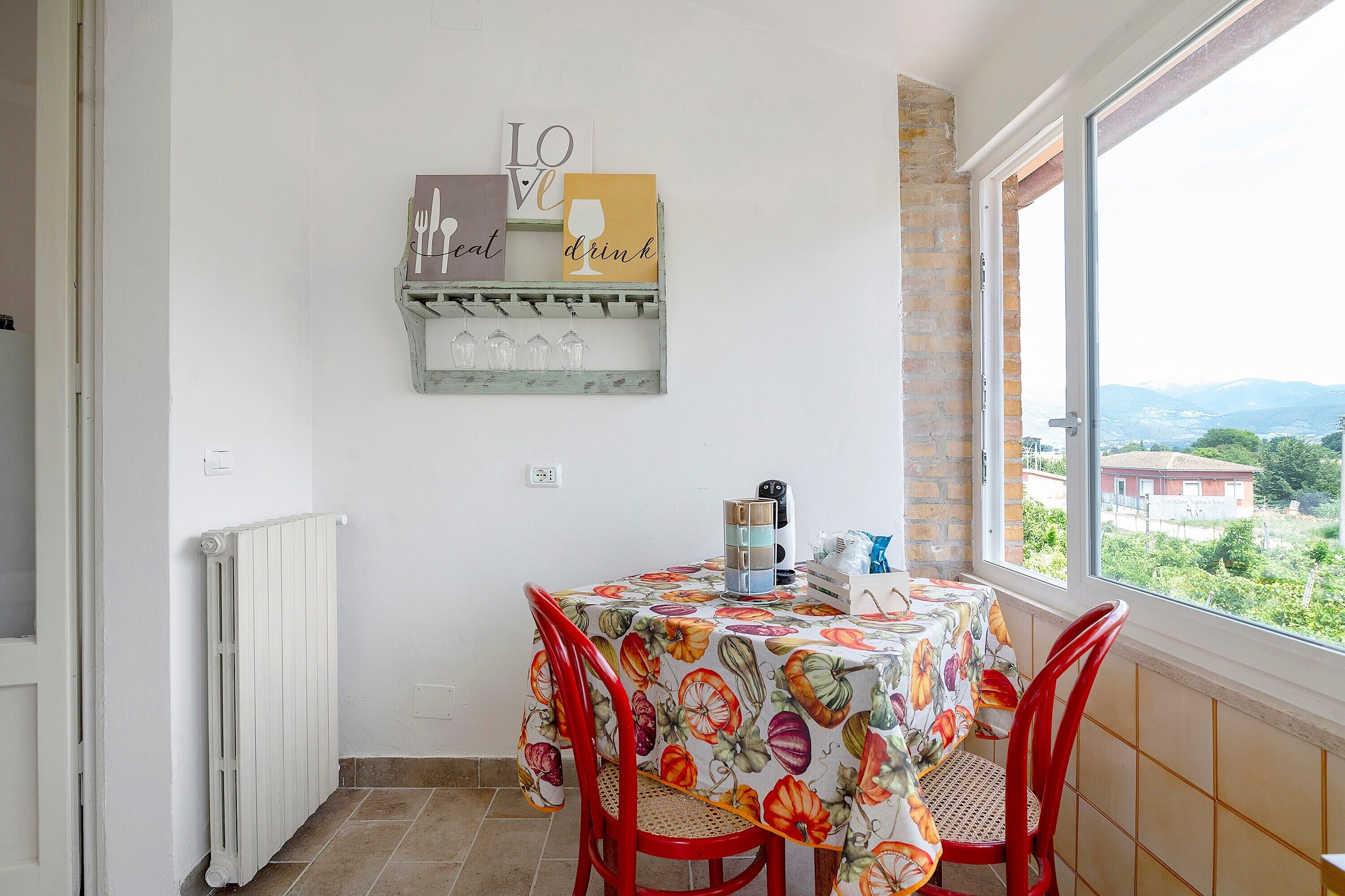 Uma área de refeições com mesa, cadeiras, radiador, prateleira na parede com objetos decorativos e uma janela com vista.