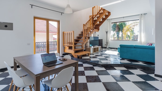 Un interior modern amb una escala de fusta, una zona de menjador amb un ordinador portàtil i un sofà blau.