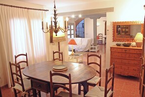 A dining area with a round wooden table, six chairs, a chandelier, a mirror, and a dresser with drawers.