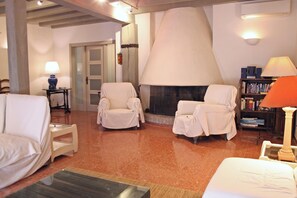 A room with a fireplace, two covered chairs, a sofa, a bookshelf, and a lamp.