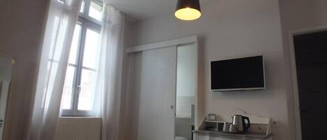 Un dormitorio con cama, una mesita de noche con hervidor, un televisor de pantalla plana, una ventana con cortinas y una puerta.
