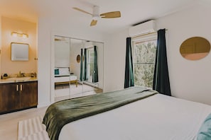 Ein Schlafzimmer mit einem Bett, einem Spiegel, einem Deckenventilator, einem Fenster mit Vorhängen und einer Wand-Klimaanlage.