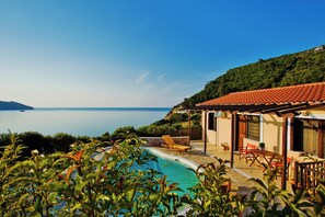 Ein Poolbereich mit Blick auf das Meer, ein Gebäude mit einem Ziegeldach und eine Hügelterrasse.