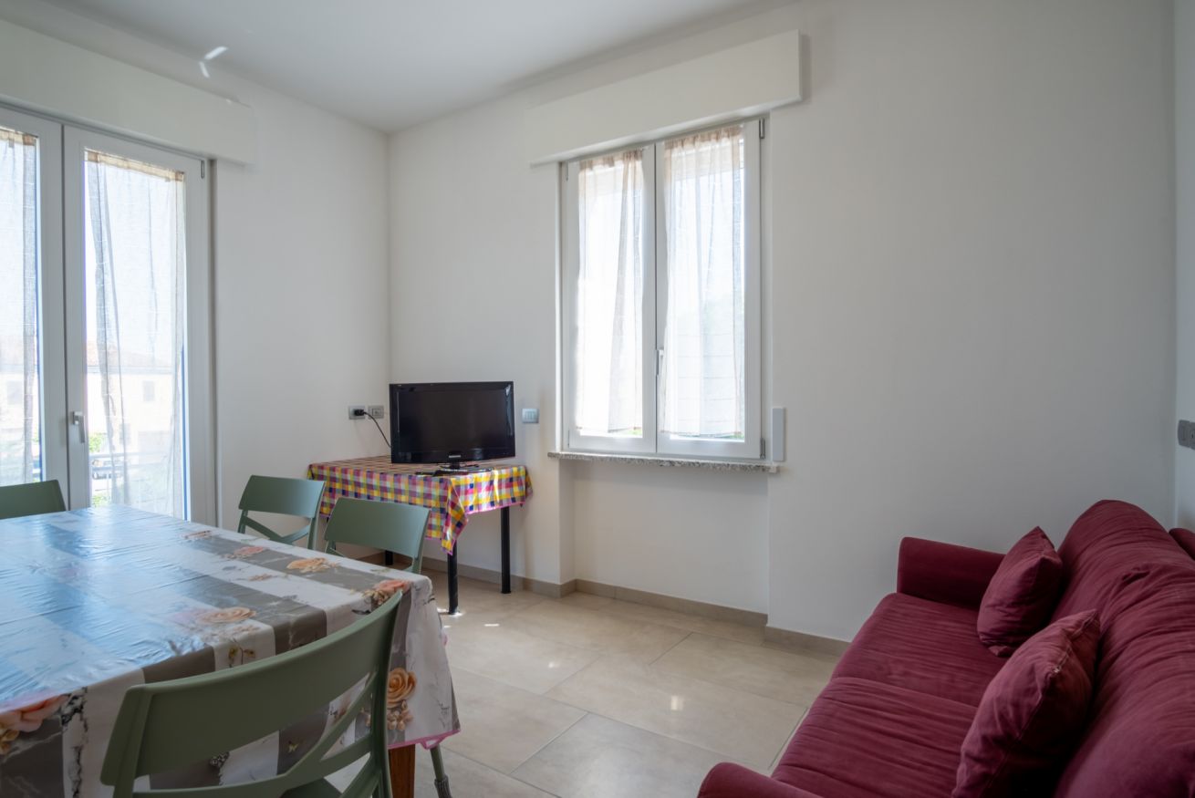 A room with a dining table, chairs, a television, a sofa, and a window with curtains.
