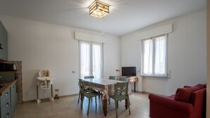 A spacious living area with a dining table, chairs, a red sofa, a television, and a kitchen area with cabinets and a chair. - GuestHost - Ezilde Residence Flat with Parking (Quarto)