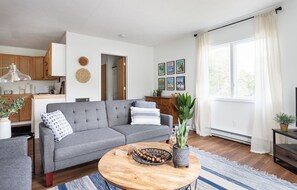 Ein modernes Wohnzimmer mit grauem Sofa, einem Holztisch und Blick ins Freie durch ein großes Fenster.
