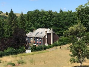Een landelijk huis met een schuin dak, omringd door weelderige begroeiing en een grasheuvel.
