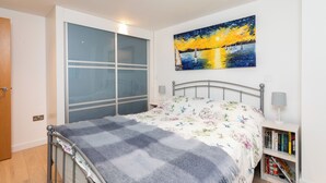 A bedroom with a bed, a nightstand with books, a painting of a sunset, and a sliding door. - Stylish Townhouse with Panoramic Sea Views | Near Beach, Canal & High Street (Hythe)