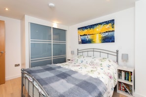 A bedroom with a bed, a nightstand with books, a painting of a sunset, and a sliding door.