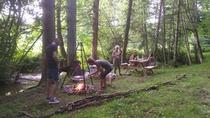 A group of people gathered around a campfire in a forested area with picnic tables and a river nearby.