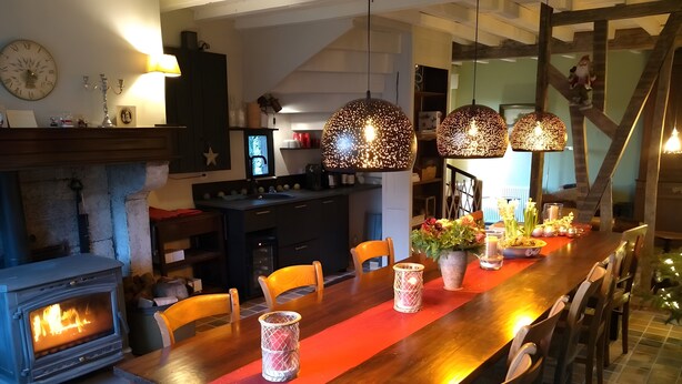 A dining area with a wooden table, chairs, a fireplace, pendant lights, and a kitchen in the background.