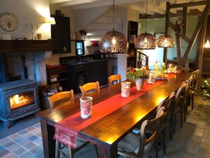 Zona pranzo con tavolo in legno, sedie, camino, lampade a sospensione e una cucina sullo sfondo.