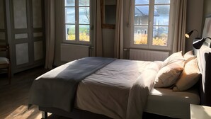 A bedroom with a bed, two windows, a radiator, a chair, and a lamp.