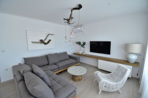 Een moderne woonkamer met een grijze hoekbank, een witte rotan fauteuil, een ronde salontafel, een flatscreen televisie en een houten plank met decoratieve objecten.