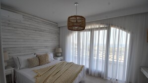 Quarto com cama, cortinas brancas, luminária pendente e vista pela janela.