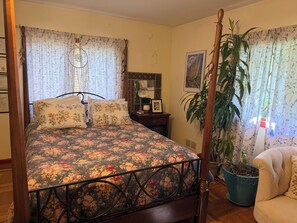 Un dormitorio con cama con dosel, ropa de cama floral, una mesita de noche, una lámpara, un cuadro enmarcado, una planta en maceta y una silla.