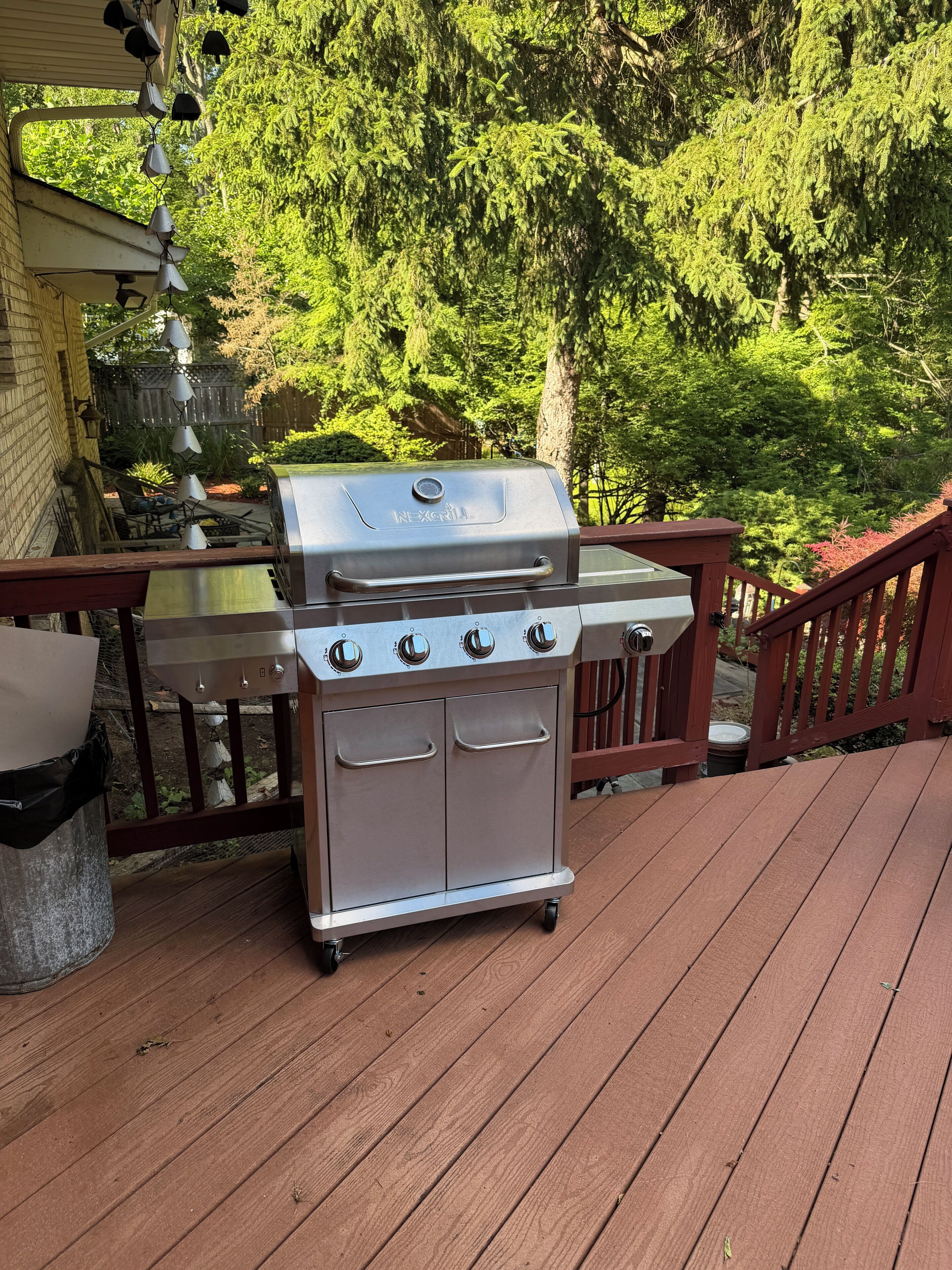 A stainless steel barbecue grill with four burners on a wooden deck, surrounded by a wooden railing and greenery.