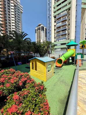 Um parquinho com escorrega e estruturas de escalada, cercado por edifícios residenciais altos.