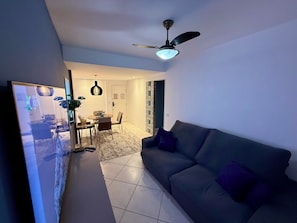 Uma sala de estar com sofá, uma área de jantar com cadeiras e mesa, um ventilador de teto e uma tela de televisão.