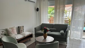 A modern living room with a grey sofa, two armchairs, a coffee table with a vase of flowers, and a view of a garden through a window. - Natura residence near salagou (Clermont-l'Hérault)