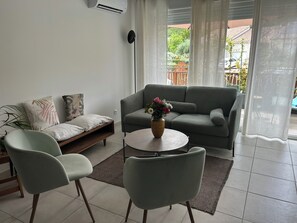 A modern living room with a grey sofa, two armchairs, a coffee table with a vase of flowers, and a view of a garden through a window. - Natura residence near salagou (Clermont-l'Hérault)
