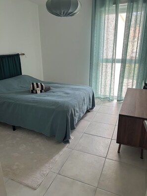 Un dormitorio con cama, una mesa de madera, un ventanal con cortinas y una lámpara colgante.