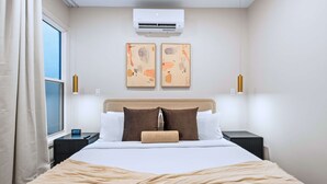 Une chambre d’hôtel comprenant un lit, une fenêtre, un climatiseur, deux œuvres d’art accrochées au mur, des lampes de chevet et une table de chevet avec une horloge.