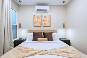 Ein Hotelzimmer mit einem Bett, einem Fenster, einer Klimaanlage, zwei Wandbildern, Nachttischlampen und einem Nachttisch mit einer Uhr.