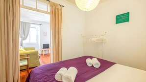 Ett hotellrum med en säng, ett lila överkast, hoprullade handdukar, en klädstång och en grön skylt på väggen, samt utsikt över ett annat rum genom en öppen dörr.