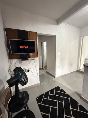 Ett rum med en väggmonterad tv som visar "NETFLIX", en golvfläkt, en svartvit mönstrad matta och en dörr som leder till ett annat rum.