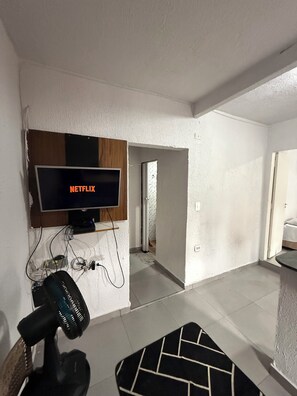 A room with a mounted TV displaying 'NETFLIX,' a ceiling fan, and a door leading to another room. - Apartamento Confortavel Mobiliado, Zona Norte SP (São Paulo)