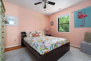 Ein Schlafzimmer mit einem Bett, einer Kommode, einem Fenster, einem Deckenventilator und einem Flamingo-Gemälde.