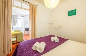 Ein Hotelzimmer mit Bett, einer violetten Tagesdecke, gerollten Handtüchern, einem Kleiderhaken und Blick auf ein weiteres Zimmer durch eine offene Tür.