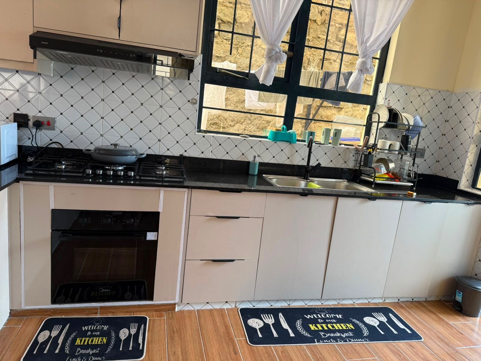 A kitchen with a stove, sink, and cabinets, a window with curtains, and a welcome mat on the floor.