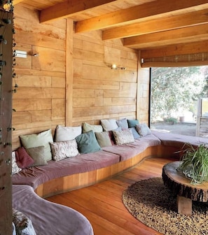A cozy wooden interior with a curved seating area, a round wooden table, and a view of greenery outside.